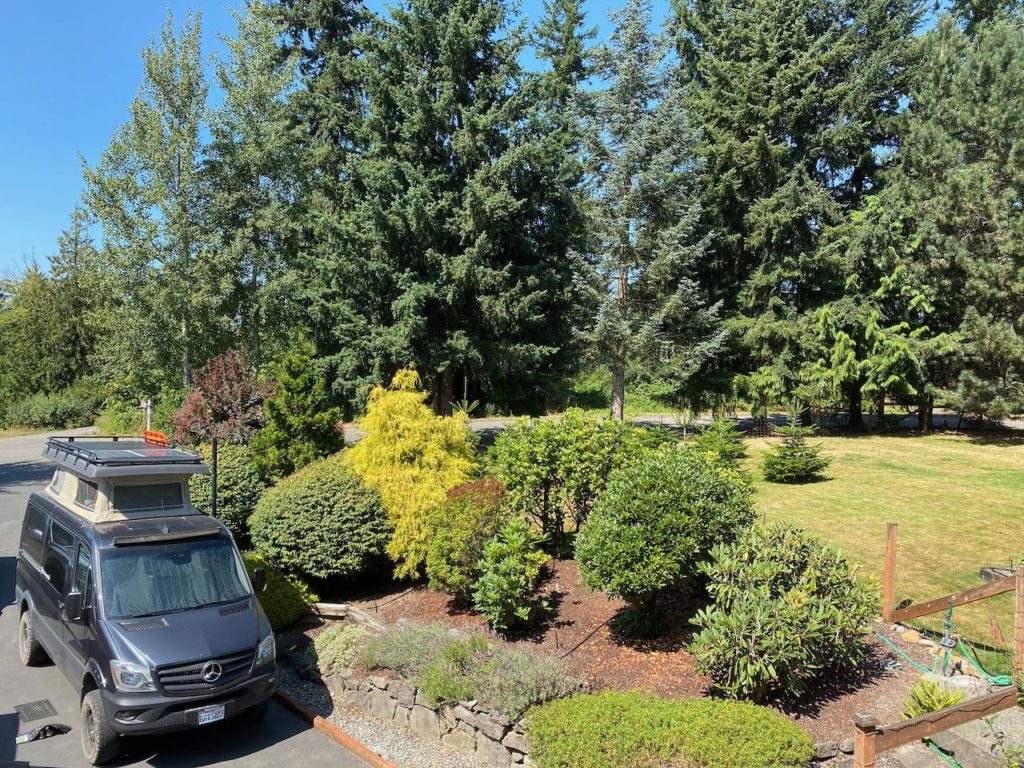 Pop top Mercedes Sprinter van parked in driveway with surrounding trees and garden. 